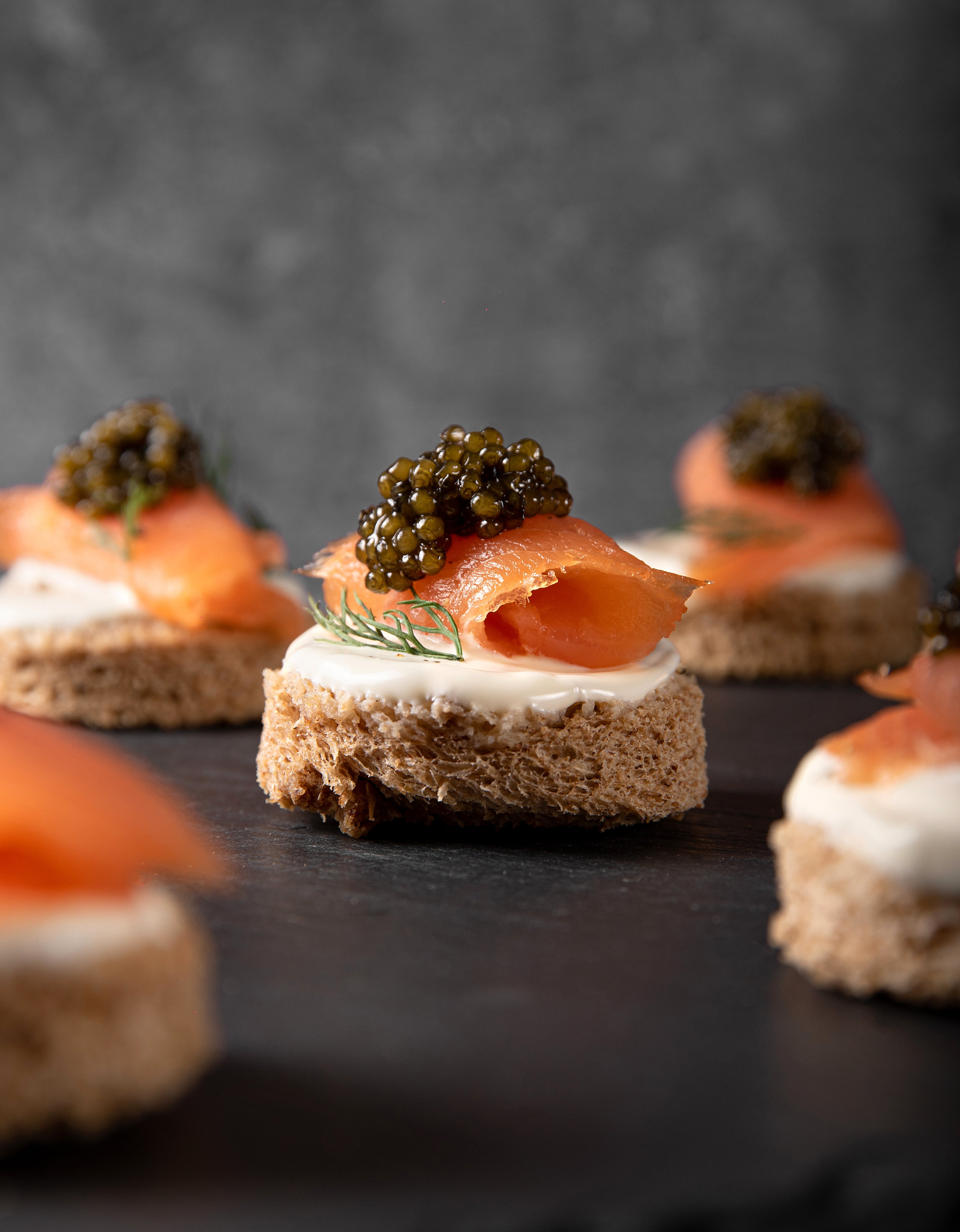 Canapés of smoked salmon, crème fraîche, and Royal Ossetra caviar with dill on round bread slices on a dark surface.