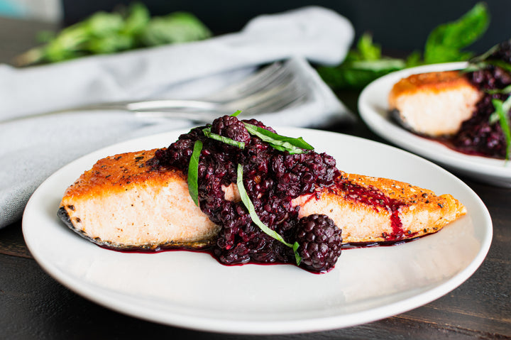 A plate containing a cut of salmon covered in fresh blackberries.