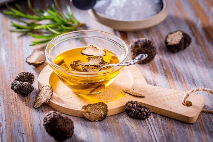 A bowl of truffle oil sits atop a round wooden cutting board with sliced truffles for garnish.