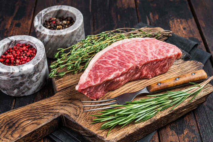 A Wagyu steak with gorgeous marbling sits on a cutting board with rosemary and other seasonings.
