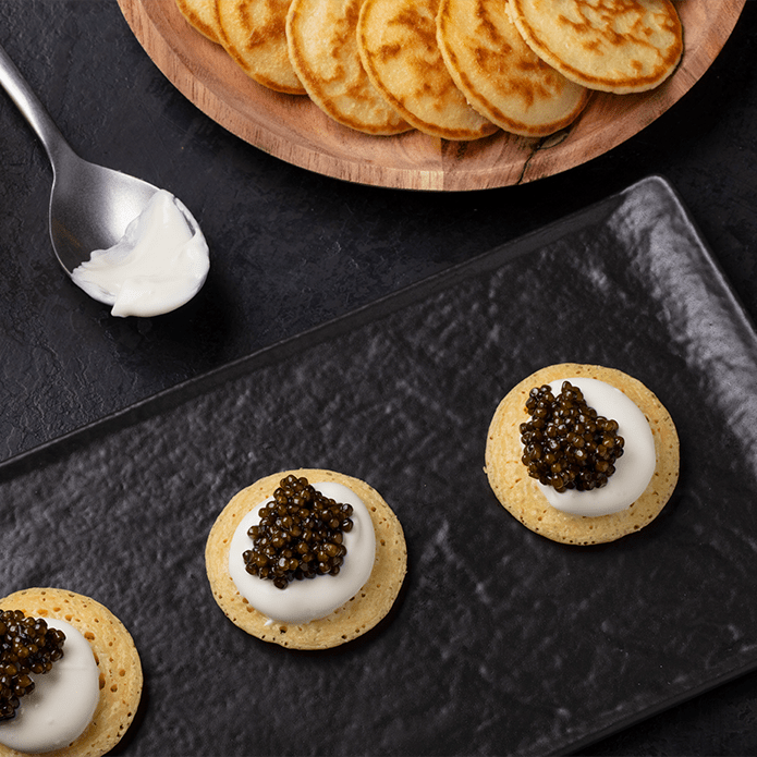 Three blinis topped with crème fraîche and caviar on a black slate plate with a spoon and a bowl of blinis behind.