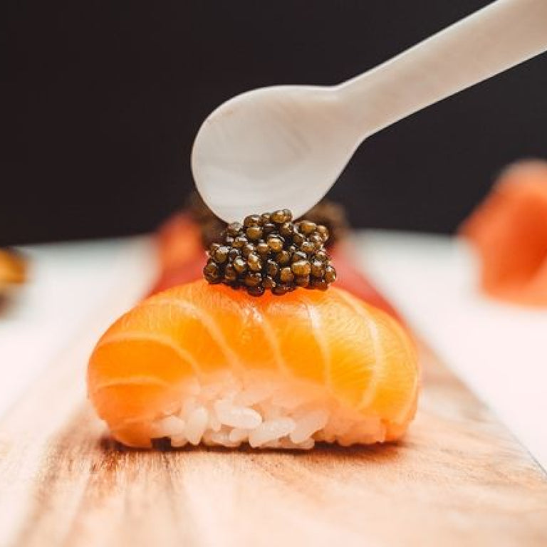 Mother of pearl spoon placing caviar on top of salmon nigiri sushi on a wooden board.