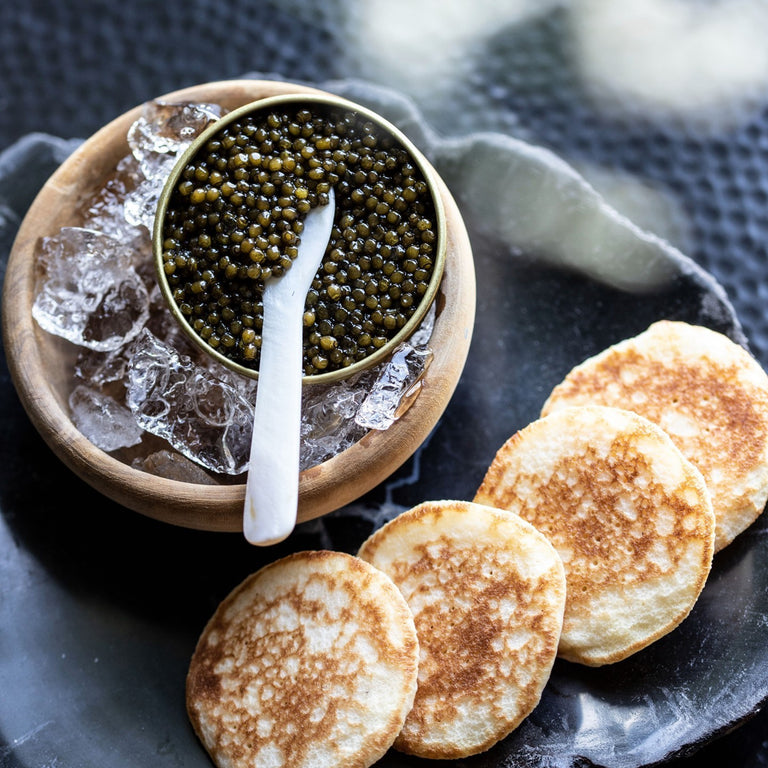 Imperia Kaluga Hybrid Reserve caviar on ice in a wooden bowl with a mother of pearl spoon, served alongside golden blinis.