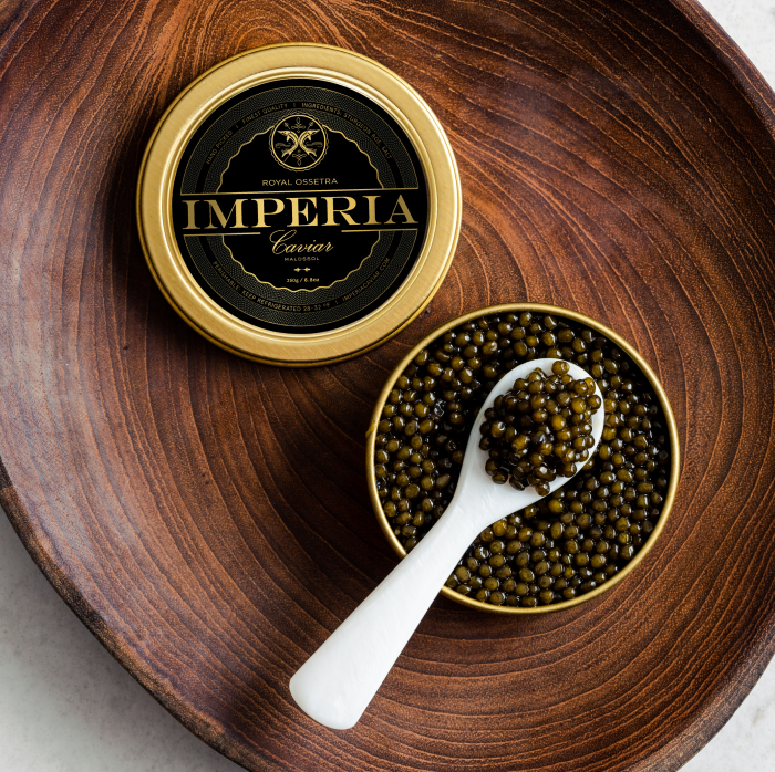 Imperia Royal Ossetra Caviar tin and open bowl of golden-green pearls with a mother of pearl spoon on a round wooden tray.