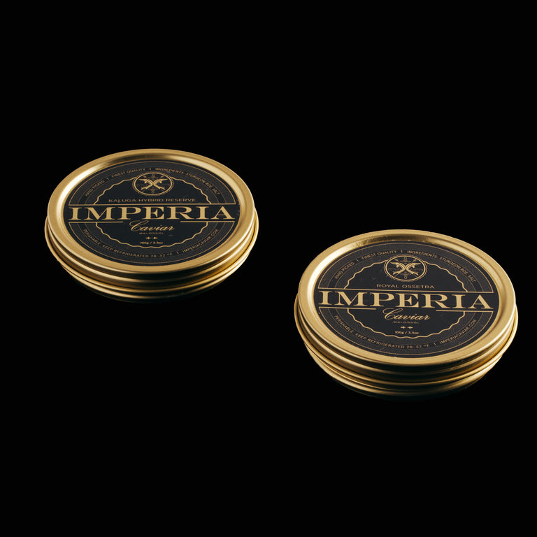 Two closed Imperia Caviar tins of Kaluga Hybrid Reserve 100g and Royal Ossetra 100g side by side on a dark surface.
