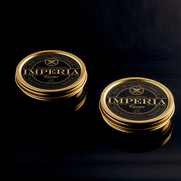Two closed Imperia tins of Kaluga Hybrid Reserve 100g and Royal Ossetra 100g side by side on a dark surface.