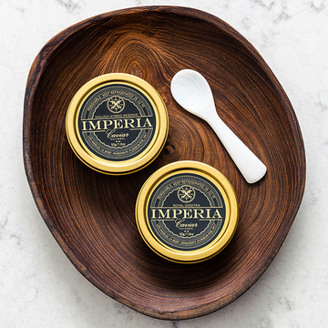 Two Imperia 50g tins of Kaluga Hybrid Reserve and Royal Ossetra with a pearl spoon in a wooden bowl on a marble surface.